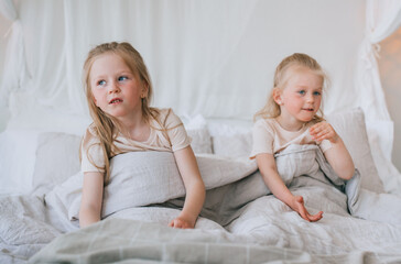 Two girls, wrapped in cozy bedding, share a playful morning in a bright, airy bedroom filled with soft textures.