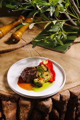 A carefully plated meat dish is presented on a white plate, accompanied by colorful peppers and broccoli. The setting features a natural wooden surface, enhancing the rustic feel.