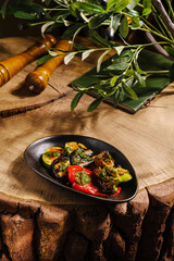 Colorful grilled vegetables arranged on a black plate sit on a rustic wooden table. Greenery and kitchen tools provide a homely, natural atmosphere enhancing the meal's freshness.