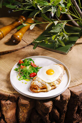 An open-faced omelet topped with a sunny-side-up egg and served with a fresh salad of tomatoes and greens is displayed on a white plate. The setting features a rustic wooden table.