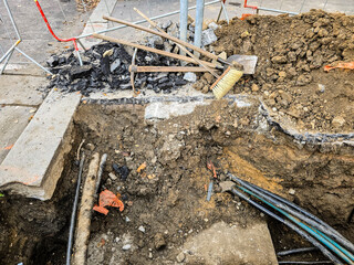 An open trench on a city street with visible underground cables and construction tools—a shovel,...