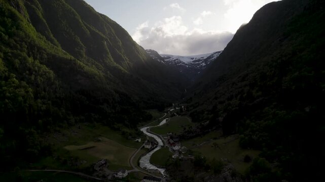 Aerial view of verdant valley and snow-capped mountains, with a winding river flowing through a village nestled below, creating a stunning contrast, Buarbreen Glacier Odda, Vestland, Norway.