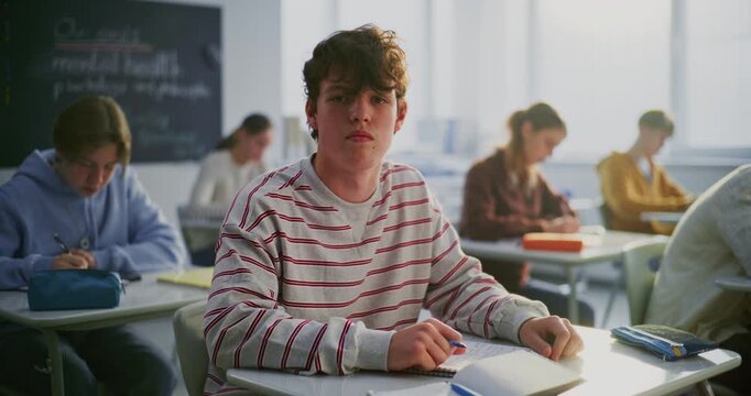 Boy in Sweater Sits at Desk With Calm, Confident Posture. Classmates Work Quietly Behind. Concept Classroom Atmosphere, Student Self Expression, Supportive Learning Environment. Slow motion. Portrait.