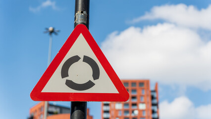 Red triangular roundabout warning sign mounted on black pole with blurred city background
