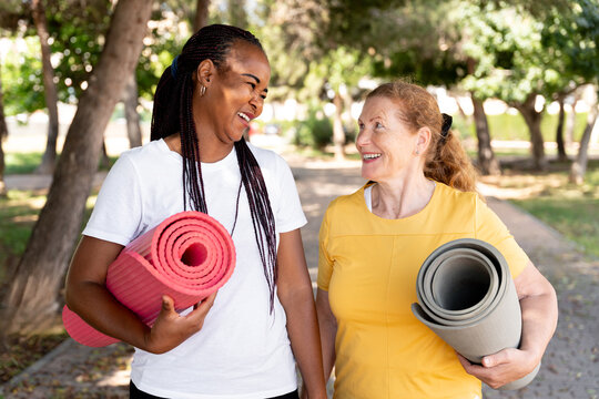 Two diverse women friends walking happily outdoors, carrying yoga mats for exercise and healthy living - Powered by Adobe
