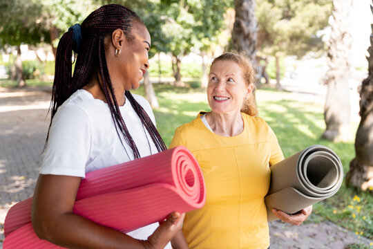 Diverse women friends carrying yoga mats, smiling, walking outdoors, enjoying fitness and wellness together