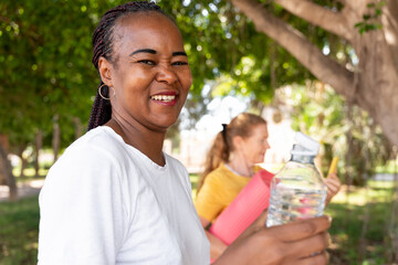 Diverse woman smiling, enjoying outdoor fitness with friends, holding refreshing water bottle for hydration