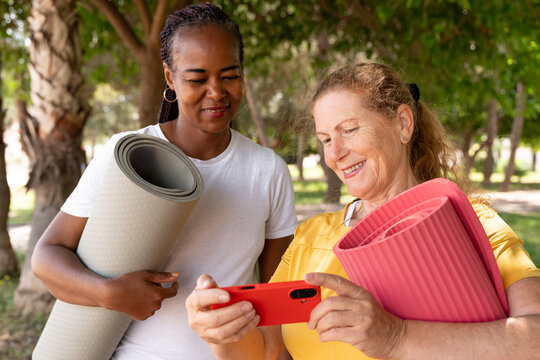 Diverse mature women carrying yoga mats, using smartphone for healthy lifestyle app in a park, smiling together