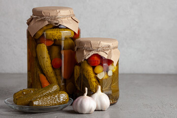 Two jars of assorted pickled vegetables wrapped in brown paper, showcasing cucumbers, tomatoes, bay leaves, garlic, and carrots. Copy space