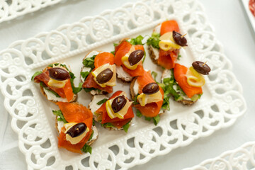 A platter of canapés featuring smoked salmon, cream cheese, and olives sits on an elegant lace tray at a cheerful gathering. The vibrant colors and fresh ingredients invite guests to indulge.