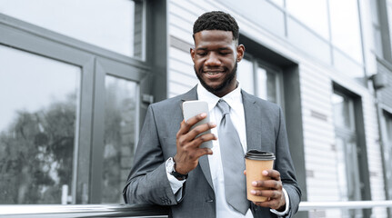 A man dressed in a gray suit enjoys his coffee while looking at his smartphone outside a contemporary office building. He appears engaged and focused on his device.