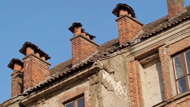 Angled view of old, brick chimneys on a deteriorating roof, against a clear blue sky