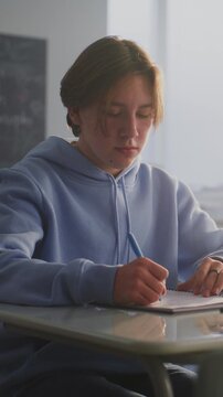 Teen Boy in Blue Hoodie Sits Alone at Desk, Lost in Thought, as Female Classmate Walking by Leaves Apple Core on Desk. Concept Teenage Loneliness, Silent Connection, Emotional Distance. Vertical Shot.