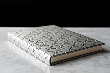 Fototapeta premium Closed silver hardcover book with embossed scale pattern on marble surface and black background, symbolizing elegance, value, and knowledge