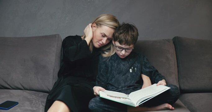 A cute schoolboy son holds a book in his hands and reads fairy tales to his mother, they sit on the couch together, a happy mother listens intently as her son reads, relaxes in the hall and enjoys rea