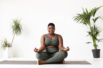 An attractive plus size black woman practices yoga in a serene home studio. Dressed in grey sportswear, she sits in lotus position on her fitness mat, smiling as she embraces the morning.