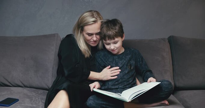 A cute schoolboy son holds a book in his hands and reads fairy tales to his mother, they sit on the couch together, a happy mother listens intently as her son reads, relaxes in the hall and enjoys rea
