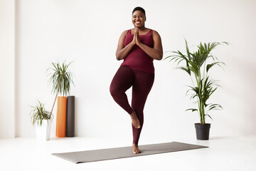 A joyful chubby young black woman performs tree pose on a yoga mat in a fitness studio. She smiles at the camera, embracing her daily yoga practice in a bright space filled with plants.