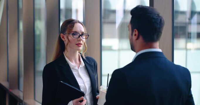 Two office workers greeting each other in hallway with friendly conversation, focus on woman holding drink