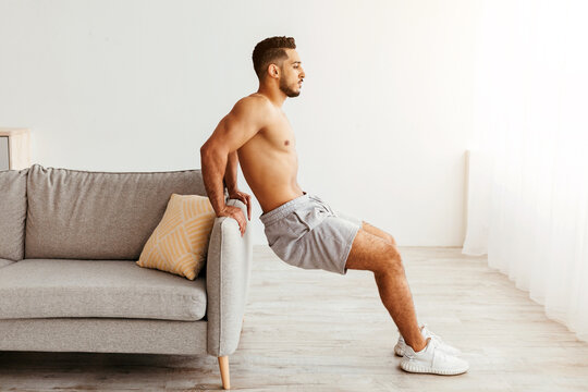 A man performs tricep dips using the armrest of a couch in his living room. Sunlight streams through the window, creating a warm atmosphere for his workout routine.