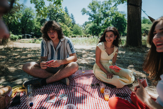 A group of friends enjoy a casual picnic on a checkered blanket, sharing cards, drinks, and laughter under bright summer sun in a park.