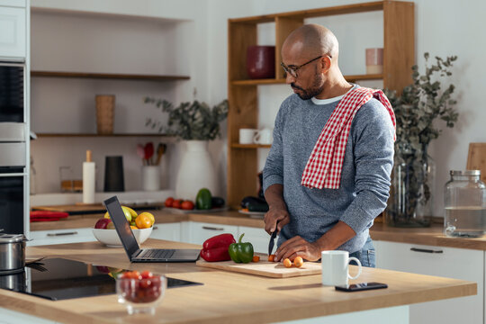 Shaved head man learning to cut vegetables through laptop in kitchen at home