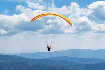 An orange and yellow paraglider soars high against a dramatic blue sky filled with white clouds, overlooking a vast panorama of layered blue and green mountain ranges.