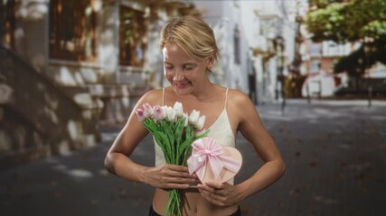 Woman with blond hair holds a bouquet of pink and white tulips and a pink heart gift box while smiling gently on a sun dappled cobbled street in front of historic buildings; romance.