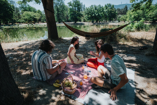 A group of friends share a relaxed lakeside picnic on a checkered blanket, sipping drinks and snacking from a basket by a calm lake surrounded by trees.