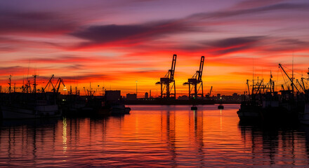 Cranes and boats silhouetted against a vibrant sunset sky reflected in the water below at the harbor