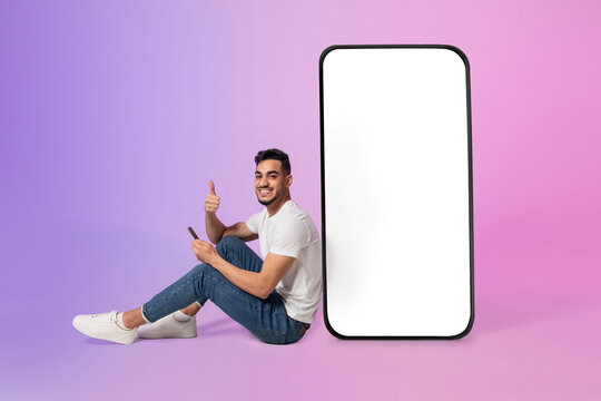A young Arab man sits comfortably with a phone in his hand, giving a thumbs up. He is next to a large cellphone with a blank white screen, surrounded by colorful neon lights.