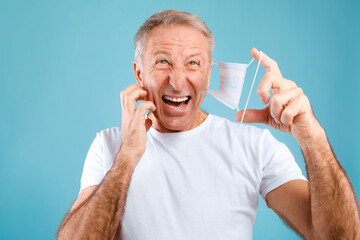 Mature man appears annoyed as he removes his protective medical mask to scratch his face. He has light gray hair and is wearing a white t-shirt against a blue background.