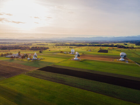Raisting, Germany - 11 November 2025: Aerial view of Raisting Earth Station's gleaming satellite dishes punctuate the lush green fields under a soft, golden sunset, creating a stark contrast.