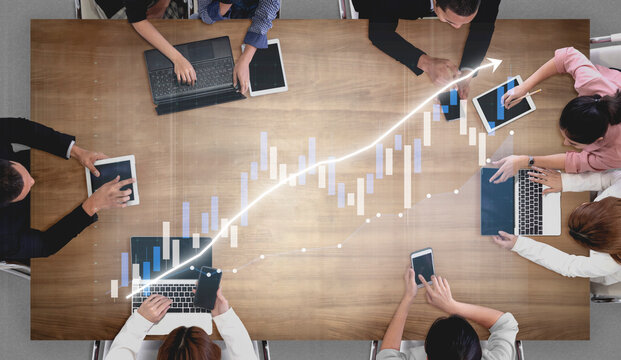 A top-down view of a business meeting showcases various professionals engaged with technology while analyzing data trends, emphasizing collaboration and innovation. Impute
