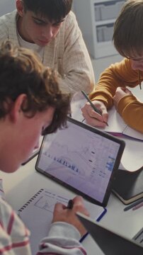 Four Young Students Gathers Around Table, Intensely Focused on Collaborative Study Session. Boys Actively Writing In Notebooks, Consulting Digital Tablet Displaying Charts and Graphs. Vertical Shot.