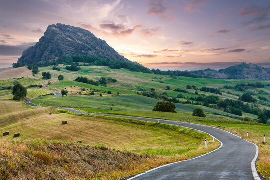 Curved country road leading to large rocky hill under colorful evening sky, Emilia-Romagna, Italy