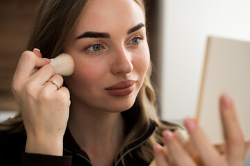 Portrait of girl applying makeup using powder brush looking in the mirror and applying cosmetic, foundation or blusher.