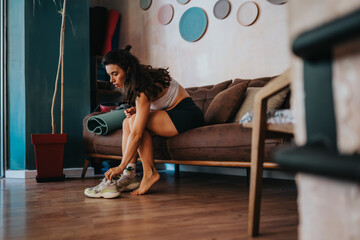 A woman sits barefoot on a brown couch in a cozy living room, tying running shoes while getting ready for a yoga or home workout. Colorful wall plates and a plant add warmth.