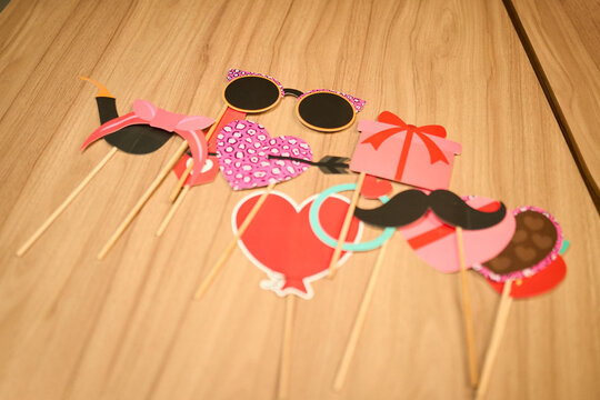 Colorful photo booth props made of paper and mounted on wooden sticks lying on a wooden table. The set includes hearts, glasses, a bow, mustache, and a gift box, designed for festive photo sessions