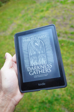 Kindle e-reader displaying the cover of the book 'While Darkness Gathers'