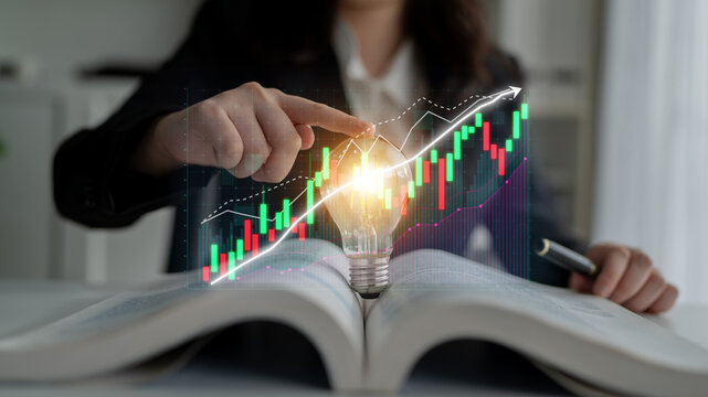 A business professional points at a glowing light bulb above an open book with a financial graph, representing the power of knowledge and innovation in strategic growth. Impute