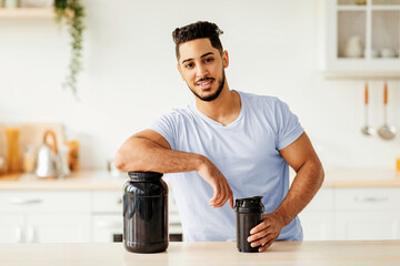 A young man stands in his bright kitchen, leaning on a large container of protein powder while holding a shaker bottle in his other hand. He smiles confidently, suggesting a healthy lifestyle.