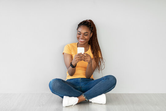 A cheerful African American woman is sitting on the floor against a grey wall, happily using her mobile phone for video chatting. She is engaged and focused on her conversation.