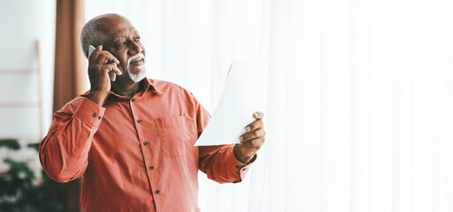 An older man wearing an orange shirt is engaged in a phone call while looking at a document. Natural light fills the room, creating a warm atmosphere.
