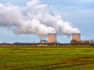 A modern power plant with two cooling towers emitting steam, visible against a backdrop of agricultural fields and a blue sky. A symbol of the contrast between industry and nature, illustrating