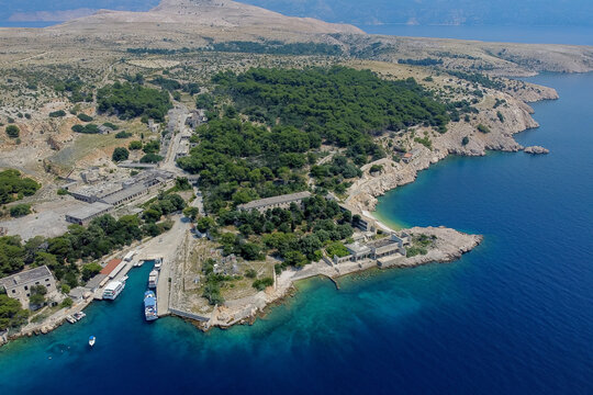 Aerial view of the stark, crumbling prison complex contrasting against the vibrant turquoise waters and dense green forests, Goli Otok, Primorje-Gorski Kotar County, Croatia.