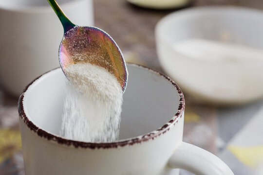 Tea spoon pouring dry milk powder into white cup. Pouring dry milk into a ceramic cup with a rainbow spoon, breakfast preparation - Powered by Adobe