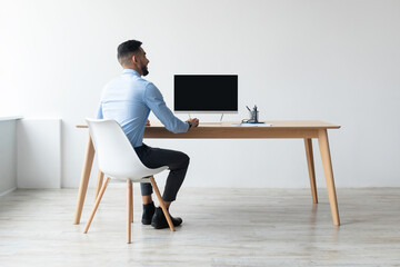 An Arab man dressed in business attire sits at a sleek desk, viewing a blank computer monitor. He appears focused, ready to engage in a webinar or video call in a professional workspace.
