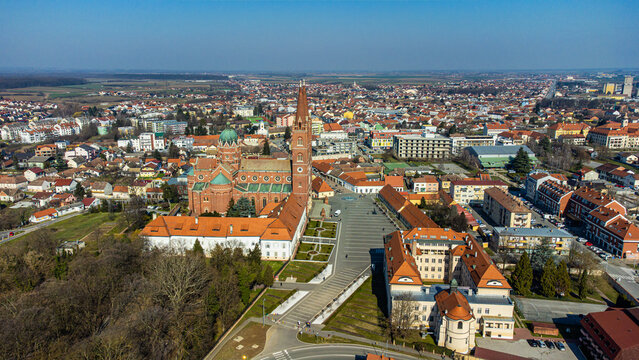 Aerial view of the magnificent Cathedral Basilica of St. Peter stands tall amidst the sprawling cityscape, its red brick contrasting against the clear blue sky, Đakovo, Osijek-Baranja County, Croatia.