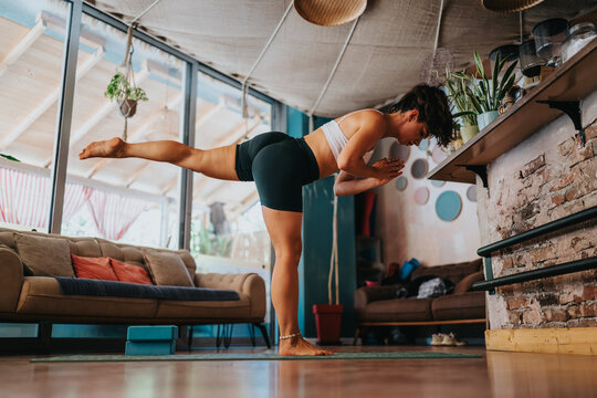 A woman in athletic wear practices a balance pose on a yoga mat in a cozy living room. Natural light, plants, and warm decor create a calm, focused fitness moment at home.